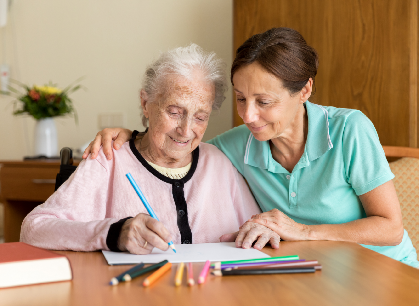 A woman and an older woman sit at a desk. The woman is helping the older woman write in a notebook.