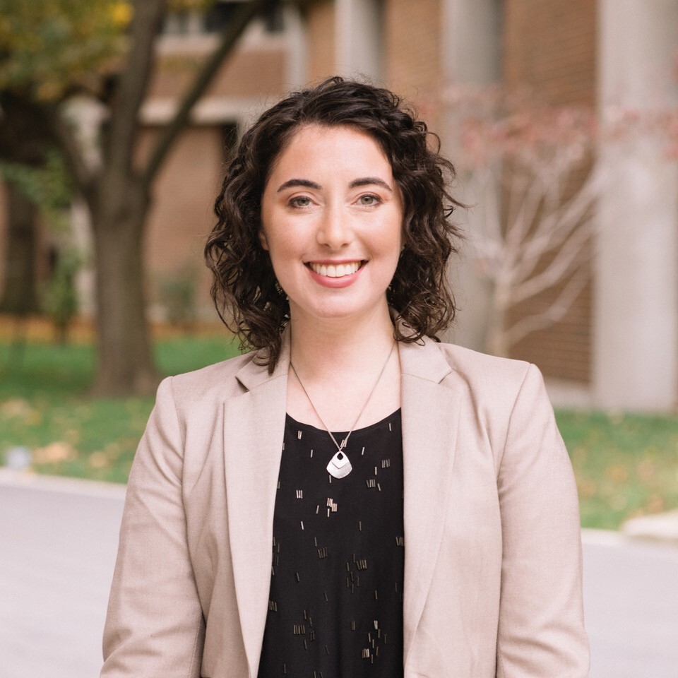 Professor Elli Weisbaum, a brown-haired woman in her thirties wearing a beige blazer and black shirt,, smiles at the viewer. A brick building and a tree are visible behind her.